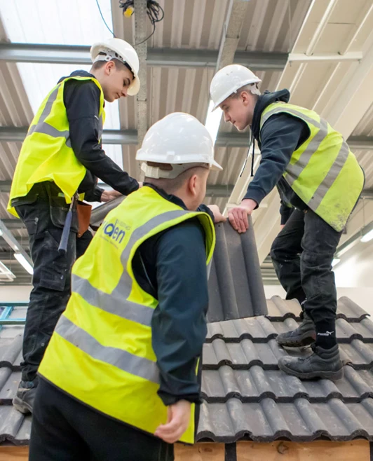 Three roofing students wearing safety helmets and high-visibility vests working together to carefully position roof tiles on a training structure Three roofing students wearing safety helmets and high-visibility vests working together to carefully position roof tiles on a training structure